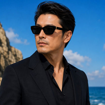 A man wearing Saint Laurent SL M153-001 Black Rectangular Sunglasses and a black blazer over a black shirt, photographed outdoors against a bright blue sky and ocean background.