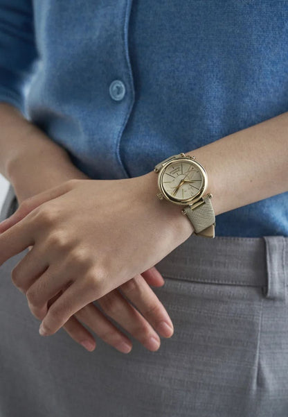 A close-up of a person's wrist wearing a Vivienne Westwood Orb Pop Champagne Gold Ladies Watch vv006gdcm with a textured beige strap, styled with a blue button-up shirt and gray trousers.